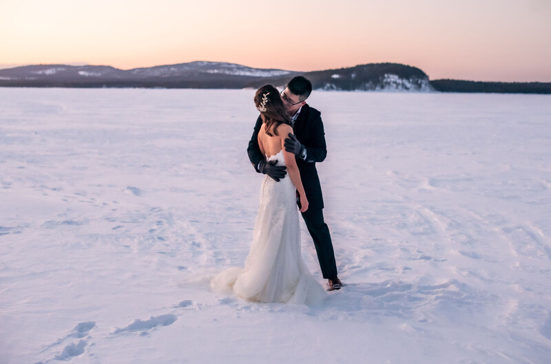 Bride and groom embracing on a frozen lake at dusk with snow-covered mountains in the background.