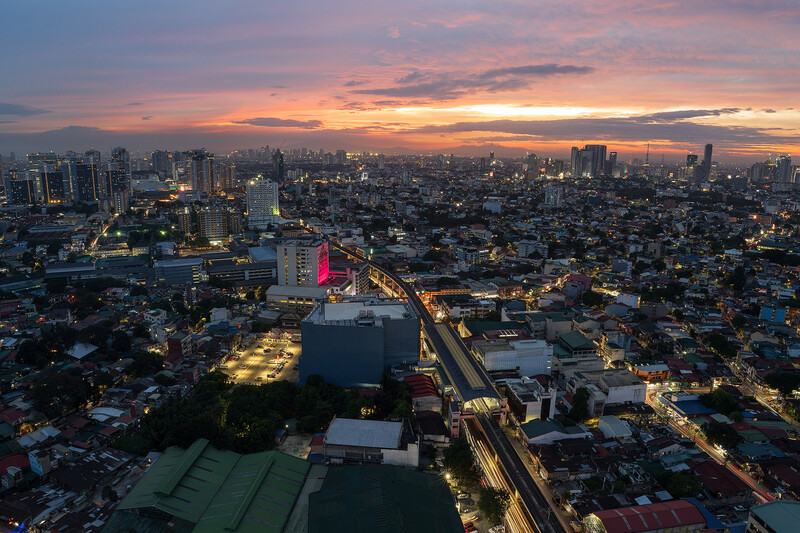 Aerial cityscape at twilight with illuminated buildings and highways stretching across a sprawling urban landscape beneath a colorful sunset sky.