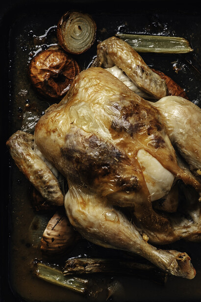 Roasted whole chicken on a dark baking tray with roasted vegetables and herbs.