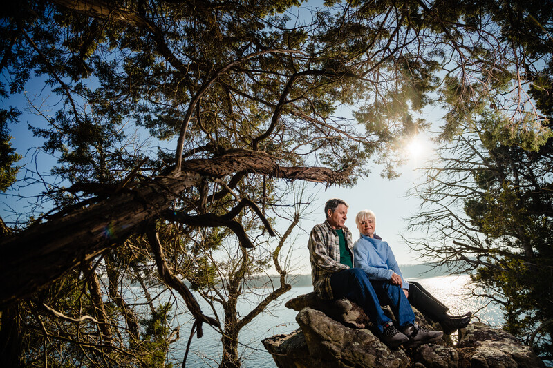Two people sitting on a rock beneath overhanging tree branches, gazing toward a calm body of water.