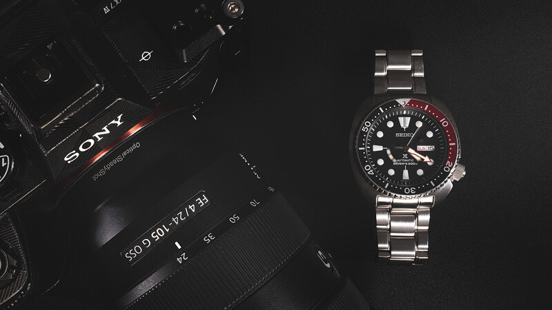 Sony camera body and lens displayed next to a stainless steel dive watch with red-black bezel on black background.
