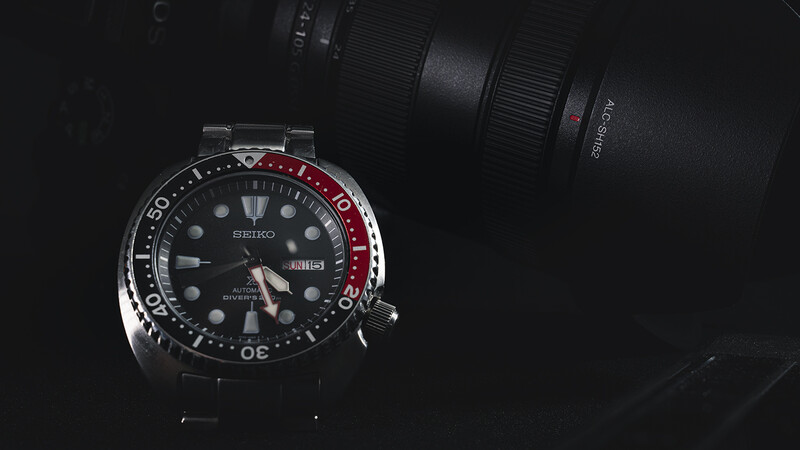 Seiko diver's watch with black dial and red/black bezel next to camera lens on dark background.
