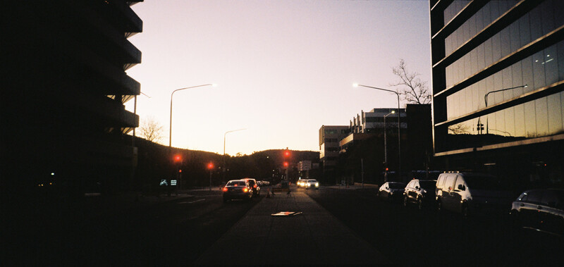 Urban street at dusk with parked cars, modern buildings, and warm street lighting creating long shadows.
