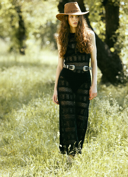 Woman in black mesh maxi dress and fedora hat standing in a sunlit grassy field.