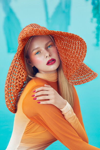Woman wearing orange textured wide-brimmed hat and matching top against turquoise background.