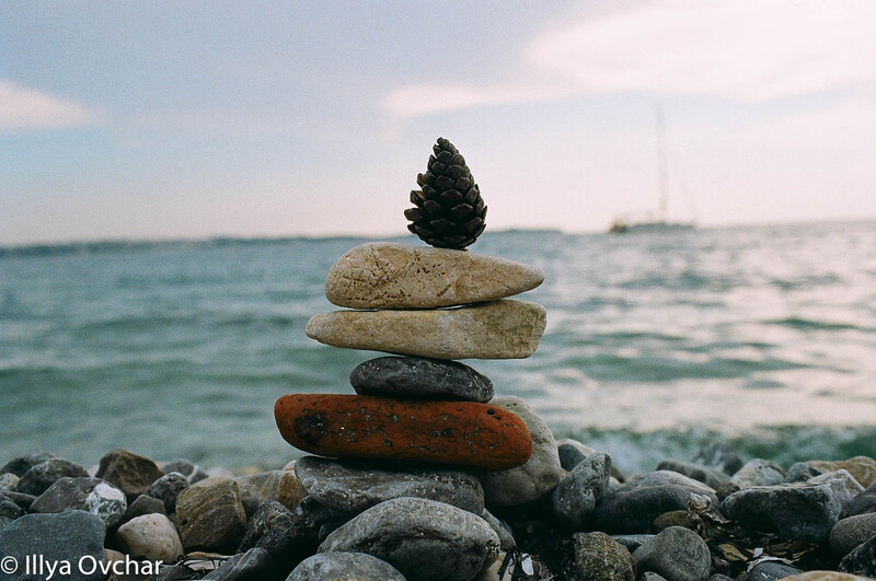 Stacked rocks and a small evergreen tree balanced on a rocky beach with calm ocean and sailboat in the background.