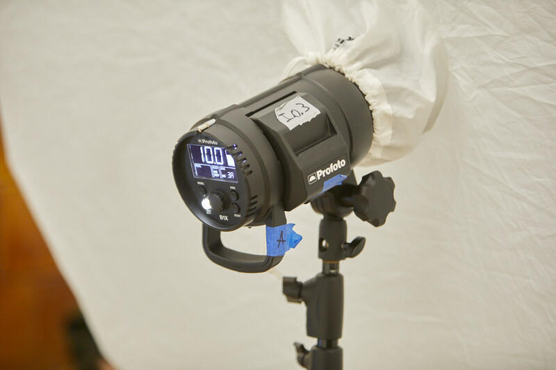 Profoto studio strobe mounted on light stand with white reflector panel visible.