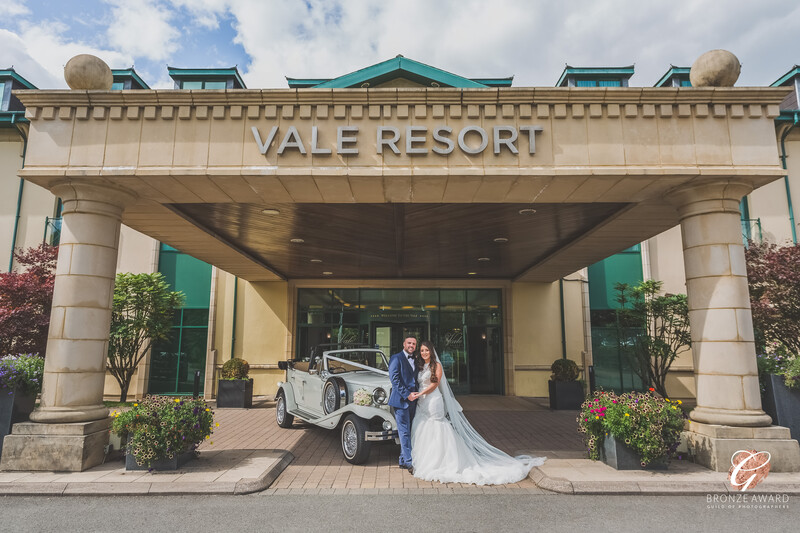Bride and groom pose with vintage white car under Vale Resort entrance canopy.