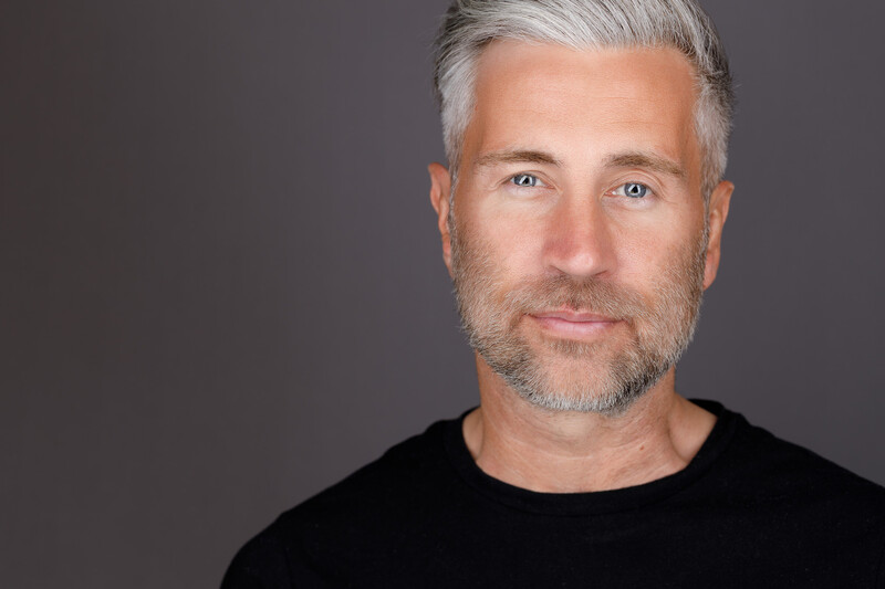 Middle-aged man with gray hair and beard wearing black shirt against neutral gray background.