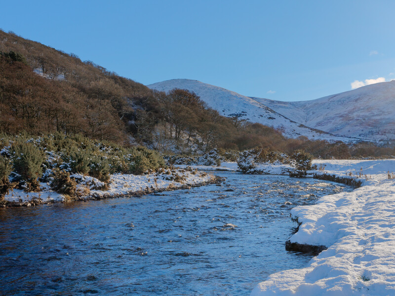 Snow-covered river valley with frost-laden trees and mountains under clear winter sky.