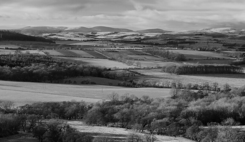 Rolling agricultural landscape with snow patches, bare trees, and mountains under overcast sky.