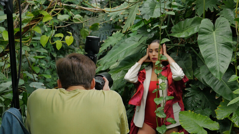 Photographer in yellow shirt capturing portrait of woman in red vest surrounded by lush green foliage.