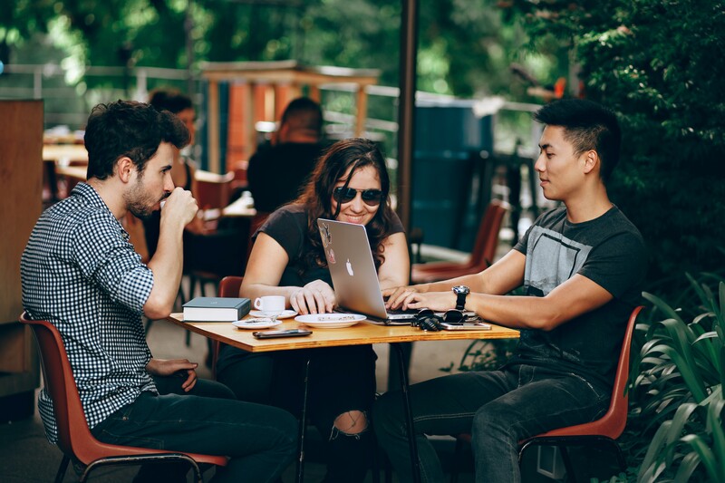 Three people collaborating at an outdoor table with a laptop and notebooks.