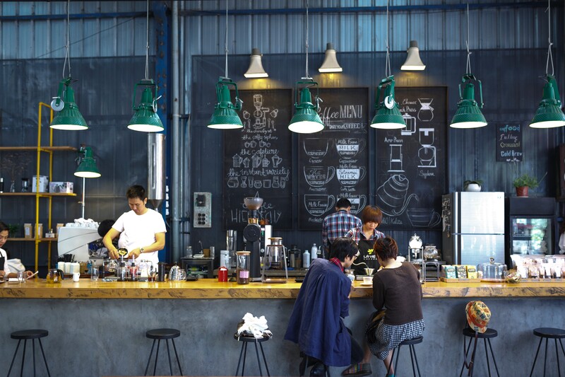 Modern coffee shop interior with green pendant lights, chalkboard menu wall, and customers at counter.