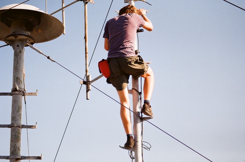 Person climbing a tall wooden utility pole against clear sky while secured by safety equipment.