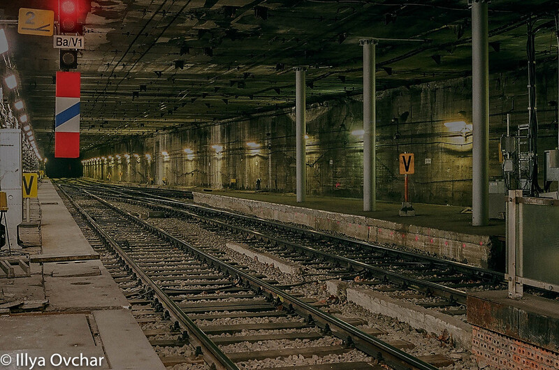 Empty subway platform with railroad tracks, concrete pillars, and red signal lights in industrial lighting.