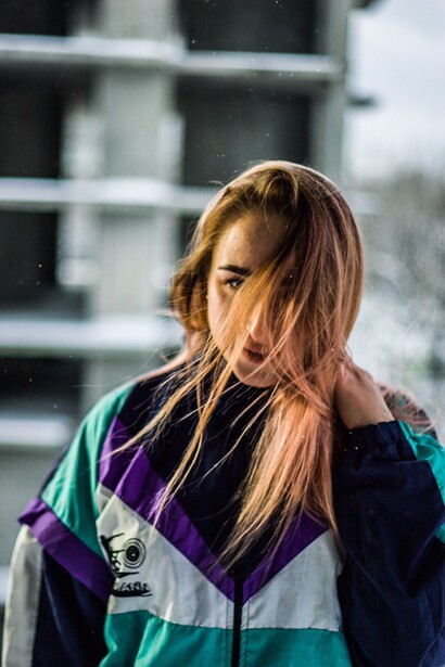Woman with long brown hair wearing a colorful windbreaker jacket, photographed outdoors with natural golden-hour light.