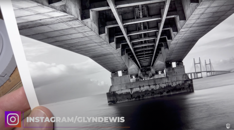 View from underneath a large bridge spanning across calm water, shot from below looking up at the structure's underside and supports.