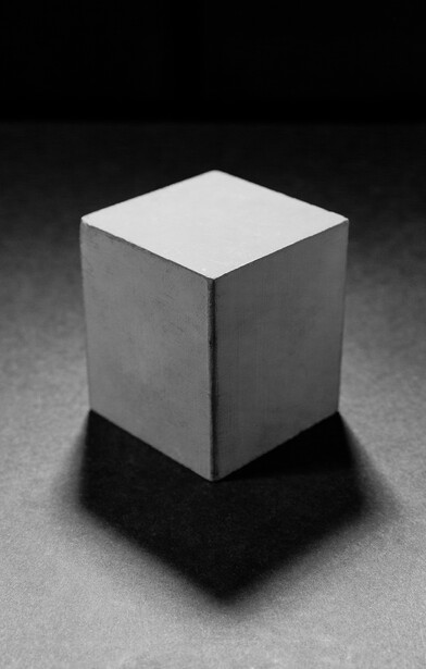 White cube casting a dark shadow on a textured gray surface against a black background.