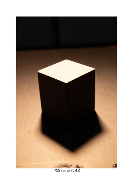 Black cube with illuminated white top surface against warm brown backdrop with dramatic shadow.