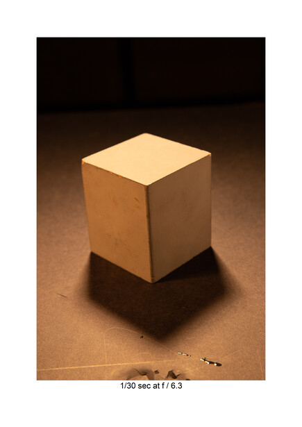 Wooden cube with warm top-lit illumination casting a sharp shadow on a textured brown surface.