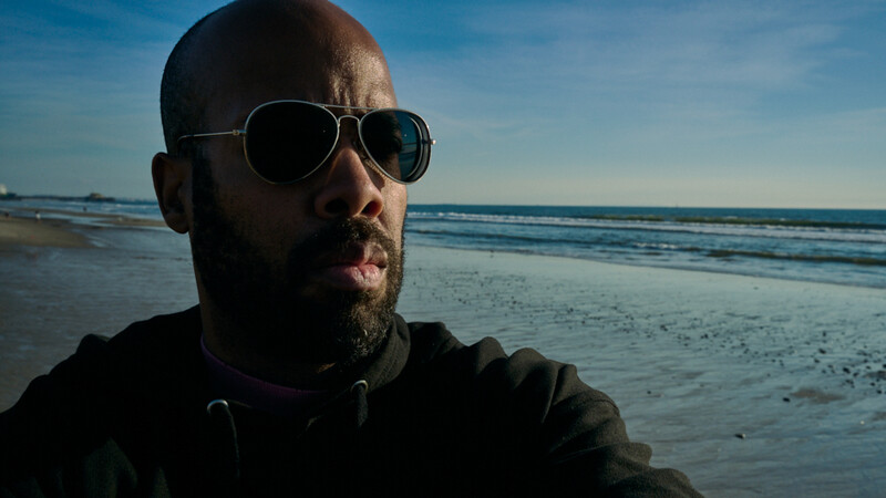 Man wearing aviator sunglasses and dark hoodie on a beach during daytime.