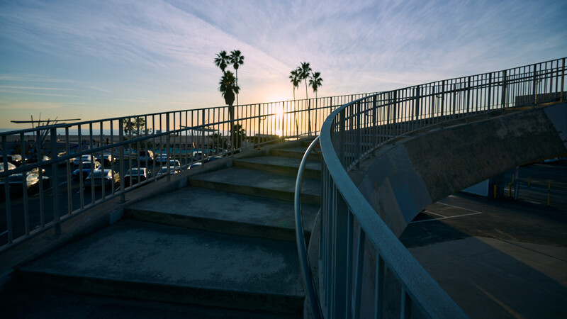 Pedestrian bridge with metal railings leading toward palm trees at sunset, with parked cars visible below.