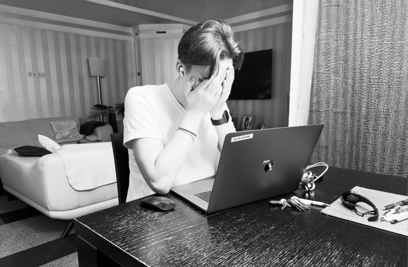 Man sitting at desk in hotel room, head in hand while working on laptop, appearing stressed or fatigued.