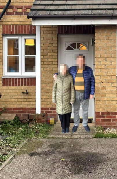 Two people standing in a doorway of a brick house, one wearing a beige gilet and the other in blue.