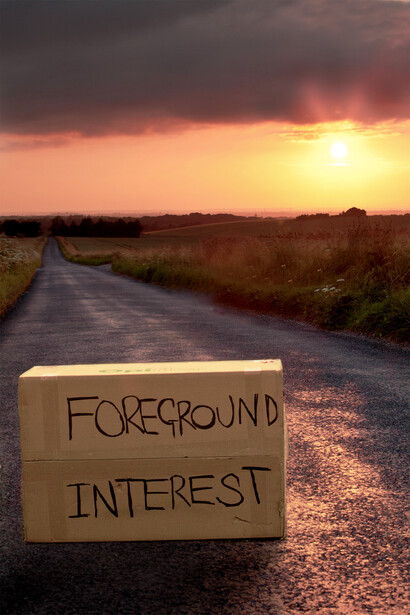 Cardboard sign reading "FOREGROUND INTEREST" placed on a wet rural road at sunset.
