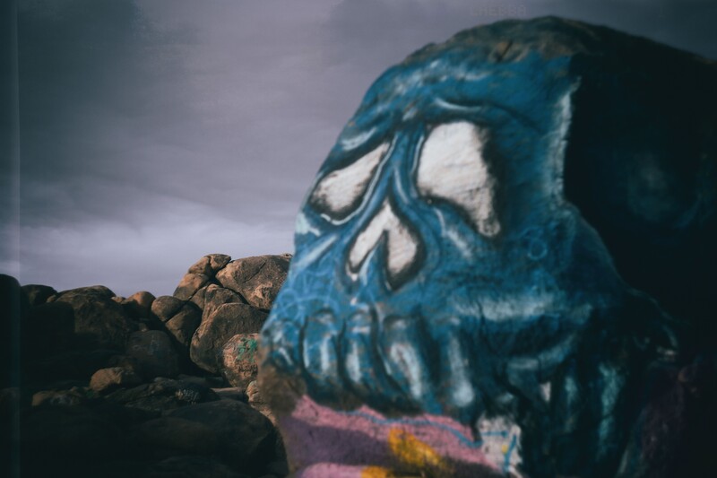 Large blue skull graffiti mural on a boulder against an overcast sky and rocky landscape.