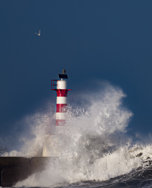Red and white striped lighthouse surrounded by powerful ocean waves and sea spray under twilight sky with crescent moon.