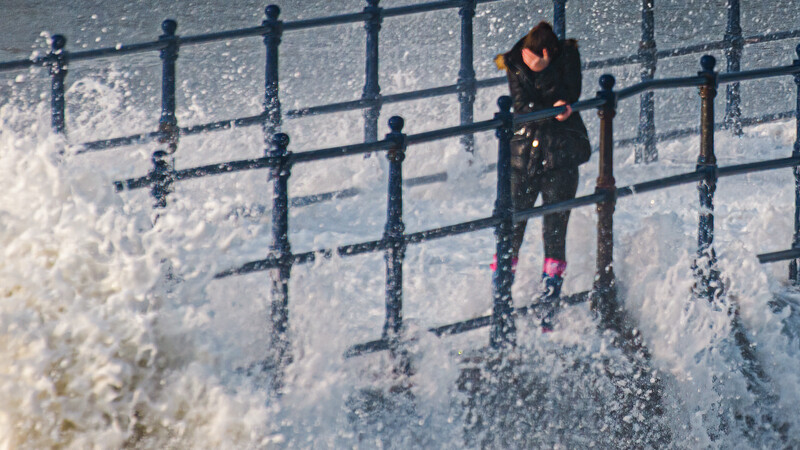 Person standing on a seaside pier railing as large waves crash against the structure.