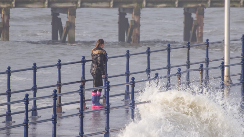 Woman and child standing at a blue seaside railing as a large wave crashes against the promenade.