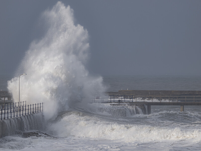 Massive ocean wave crashes against a beach fence during stormy conditions.