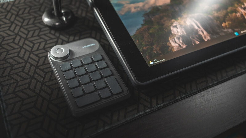 Compact keyboard controller with grid of keys and rotary dial next to tablet displaying landscape photography.
