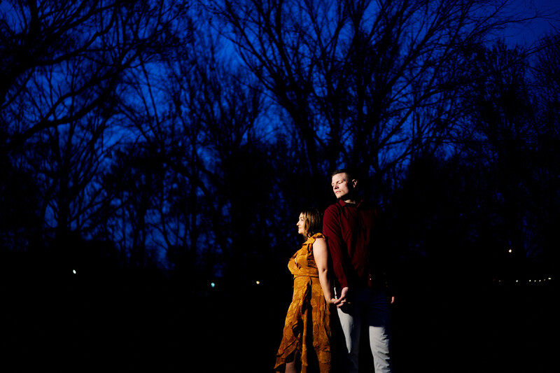 Two people standing together at night under bare trees with blue ambient lighting.