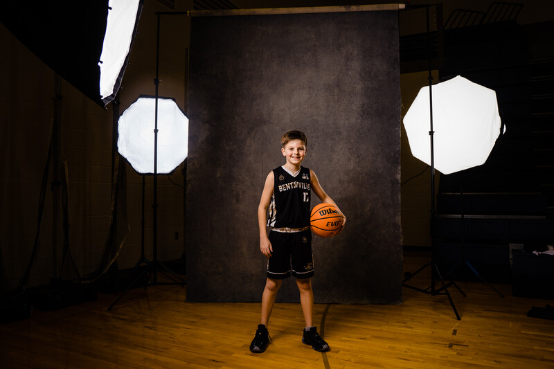 Young athlete holding a basketball in a studio lit with large octagonal softboxes and a dark textured backdrop.