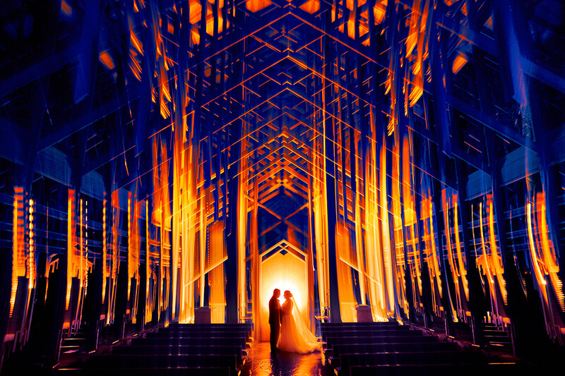 Two silhouetted figures stand in a cathedral-like architectural passage illuminated by warm orange and yellow uplighting against deep blue shadows.