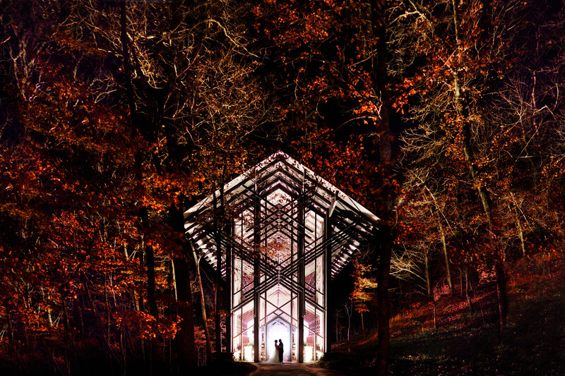 Geometric wooden structure silhouetted against vibrant autumn foliage and a glowing interior light.