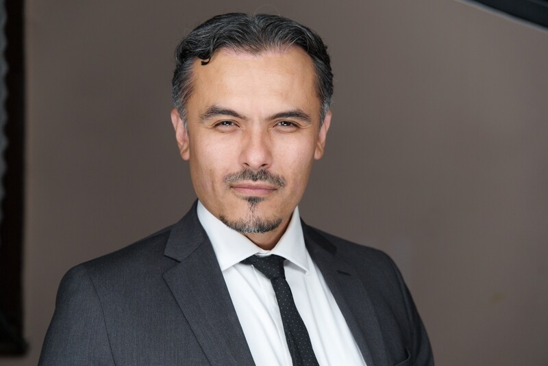 Professional headshot of a man in dark business suit and tie against neutral gray background.