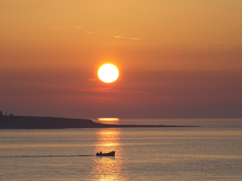 Fishing boat silhouetted on calm water beneath a large orange sun at sunset.