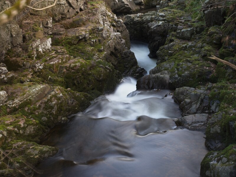 Cascading stream flowing through moss-covered rocky gorge with long exposure blur effect.