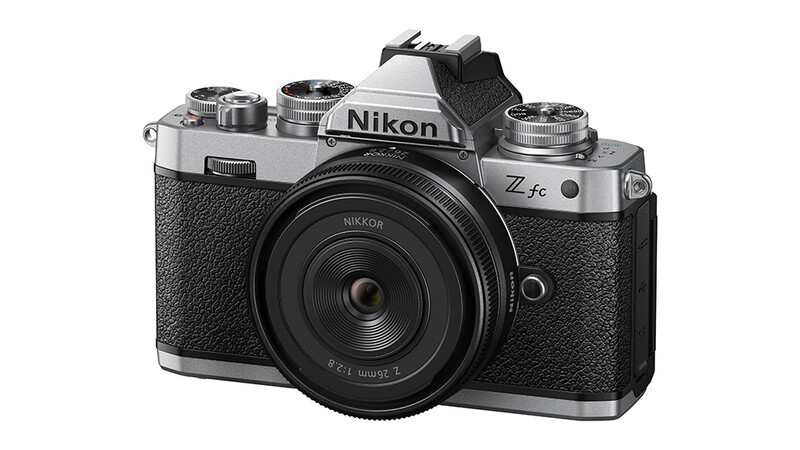 Nikon Z fc mirrorless camera with silver top plate and black body, shown from front with standard zoom lens attached.