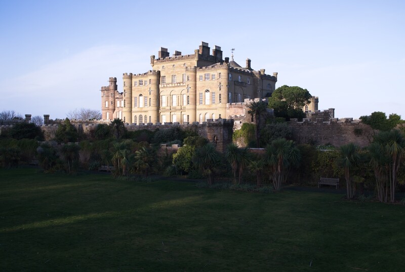 Golden-stone castle on a hilltop surrounded by green fields and trees under clear sky.