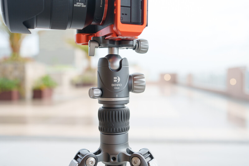 Red camera rig mounted on ball head atop tripod legs with blurred rooftop background.
