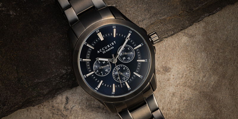 Black chronograph watch with three subdials on a textured gray concrete surface.