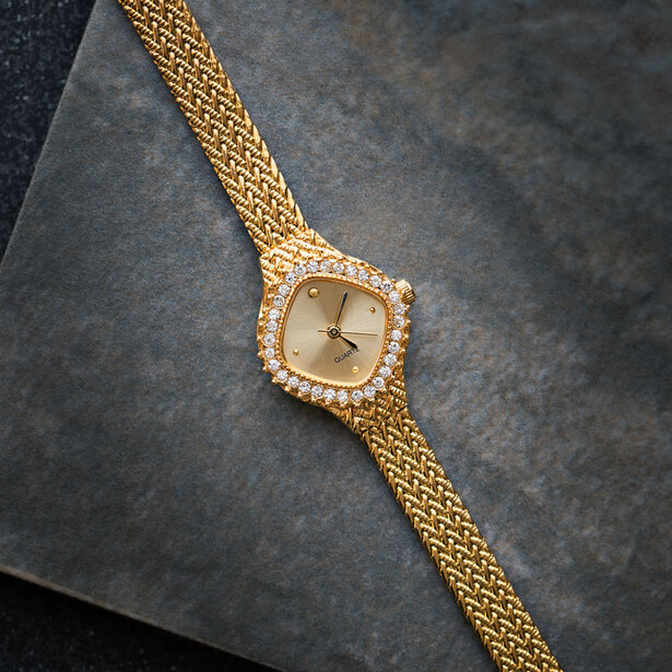 Gold mesh band watch with diamond-set square face on dark textured background.