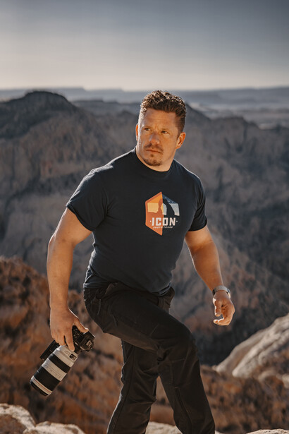 Male photographer holding camera gear at mountain overlook during golden hour.