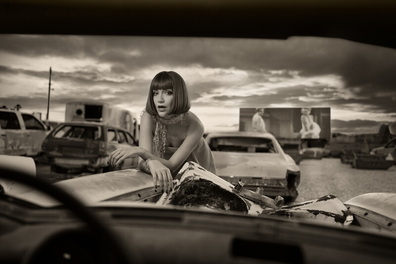 Woman in patterned dress posing on abandoned car in junkyard under dramatic cloudy sky.